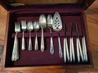 Full shot of the flatware set neatly arranged in a worn solid wood case with inset velvet lining, showing the forks, spoons, knives, and serving pieces.