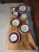 Full set of Sebring Pottery Co burgundy gold trim dinnerware with plates and bowls arranged on a wooden table near leather furniture.