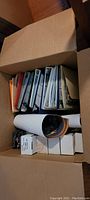 Photo of a box containing multiple binders filled with trading cards, rolled posters, and white boxes, showing organization and quantity of items.