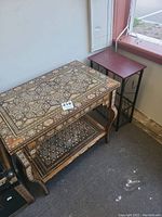 Two side tables shown together near a window. One highly detailed inlaid wooden table with drawer and lower shelf, and the other simpler table with reddish wood top and metal legs.