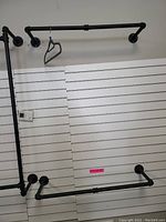 Full view of the largest metal pipe clothes rack mounted on a white slat wall panel with screw flange mounts. One hanger present on the rack.