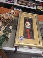 Framed decorative clown mask with a white face and red tassel on a display table.