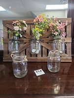 Front view of five glass jars on wooden frame; three jars contain artificial flowers and string lights, two jars empty at base
