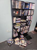 A black media shelf loaded with DVDs, Blu-rays, VHS tapes, and additional stacks of media on the floor alongside a black DVD player with cables.