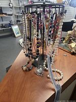 Black metal jewelry display rack with numerous necklaces hanging, including faux pearl strands, colorful beads, and heart pendant necklace.
