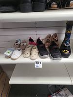 Seven pairs of kids and toddler shoes displayed on shelf including baby booties, sneakers, sandals, and rain boots.