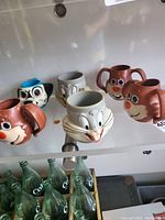 Six vintage Looney Tunes character ceramic cups, displayed on a glass shelf showing various characters' faces and ears as handles.