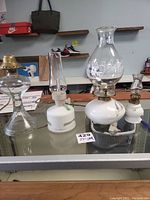 Four oil lamps arranged on glass countertop: one with clear glass base and brass burner, two white ceramic base with floral designs, and one larger white ceramic base with etched glass chimney showing dancing figures.