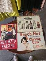 Two vintage advertising signs displayed on a table with clothes in background. One sign is Coca-Cola bottle evolution, the other is Beech-Nut Chewing Gum.
