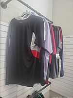 Photo of men's tops on hangers showing a black t-shirt with side panels, checkered shirt, and various t-shirts in grey, red, and blue tones.