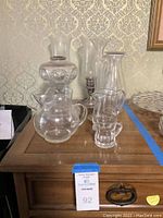 Photo showing various glass pitchers and glass lamp shades arranged on a wooden surface with a wallpaper background.