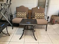 Two matching outdoor metal frame chairs with beige cushions and patterned pillows positioned behind a square dark metal table with decorative lattice top on tiled floor.