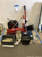 Wide view of all car repair tools including hydraulic jack, battery charger, compressor, cables, lug wrenches, and lubricant spray.