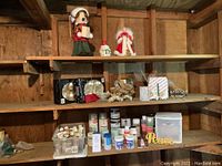 Photo showing upper and middle shelves with Christmas ornaments including snowman, angel, dove figures, and multiple craft paint bottles and containers on the lower shelf.