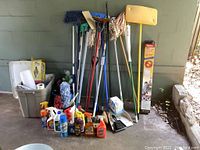 Photo showing mop handles, spray bottles, dustpan, and cleaning supplies organized on ground against a wall.