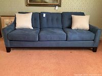 Front view of blue couch with two cream pillows on each side, set against patterned wallpaper and carpeted floor.