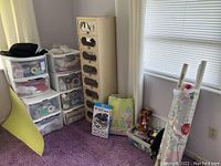 Stacked plastic storage drawers containing ribbons, gift wrapping supplies, and craft materials in a room corner with a purple carpet and a window.
