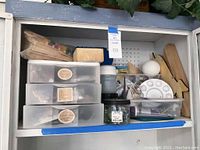Photo showing storage cabinet with plastic stacking drawers containing craft supplies including labeled containers for pebbles, small nails, paint bottles, glue, and other materials