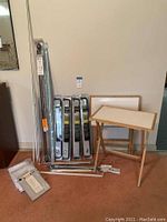 Photo showing four matching adjustable metal curtain rods, two wooden TV trays folded, and additional curtain rods and hardware packed near the wall.