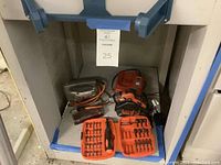 Photo shows Black & Decker electric jigsaw with orange and black body, charger, and accessories on floor inside garage space.