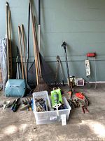 Full overview of long-handled rakes, sprinkler parts in plastic bin, some hand tools and grass trimmer