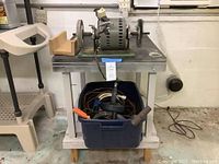 Photo showing grinding wheel attached to metal bench with an attached wooden miter box and storage container with tools below.