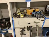 View of shelf and lower shelf with clamps, tape measure, yellow toolbox, stapler, elastic straps, cooler and other items.