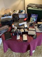 View of entire lot with electronics and office supplies on a table, includes laptop, cassette devices, razors, a mouse, staplers, and other miscellaneous items.