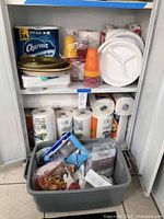 Photo of shelves containing 24 mega rolls of Charmin Ultra Soft toilet paper, stacked packs of colorful disposable plastic cups, disposable white divided plates, and paper napkins on the top shelf, with various other paper products below.