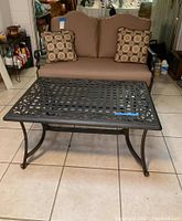 Loveseat sofa with dark metal frame, brown cushions and two patterned pillows with patio table in front.