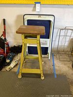 Side view of yellow metal-legged stool with wooden seat and folding platform cart behind it