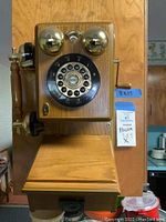 Front view of the reproduction antique wall phone showing the rotary style dial with push button numbers, two silver bells, wooden receiver handle, and part of the wooden back plate with tag indicating size 8x17.
