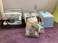 Brother sewing machine in brown carrying case with power cord and foot pedal. Multiple plastic containers with ribbons and craft supplies, hot glue guns in packaging.