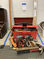 Overall view of red plastic toolbox filled with various hand tools including hammers, pliers, and saws arranged in front of the box.