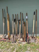 Full view of assorted gardening tools including rakes, shovels, axe, pruning shears, and sledgehammer lined up against a wall on grass.