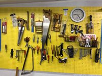 Wide view of yellow pegboard wall with assorted hand tools pinned including handsaws, pliers, scissors, clamps, and other hardware items