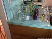Wide view of all glassware items including vases, covered cake stand, and dispensers on blue counter