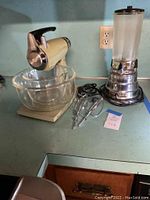 Wide shot of vintage Sunbeam MixMaster stand mixer with large glass bowl, two beaters and power cord next to vintage Waring blender with ribbed glass jar and chrome base on kitchen counter.