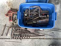 Top view of blue bin filled with vintage rusty hand tools including hand braces, clamps, and drill bits spread around on the ground.