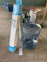 Photo of large clear water jug with attached white hand pump, in a garage setting with other household items in background.