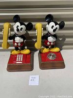 Two vintage Mickey Mouse telephones shown side by side, one with rotary dial and one with touch tone keypad, both with yellow handset and Mickey Mouse figure.