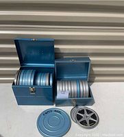 Two blue metal film reel cases opened showing multiple metal 8mm film reels inside, with a few reels outside including one labeled empty and one containing film.
