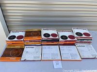 Stacked assortment of approximately 35 vintage reel to reel audio tapes in original packaging, featuring Ampex and Ambron brands, with printed inserts visible.