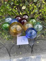 Eleven colorful glass balls arranged together on a glass table outdoors with greenery in background.
