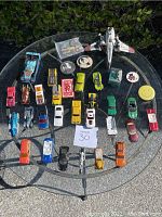 All toy vehicles arranged on circular glass table outdoors, showing assortment of Hot Wheels diecast cars, trucks, planes, and collectible pins.