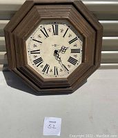 Hexagon shaped wooden framed clock with Roman numeral face and black ornate clock hands on a white dial. Clock is battery powered and untested.