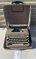 Photo shows gray Royal Quiet De Luxe typewriter inside original hard shell carrying case, lid open, with black handle on top and chrome trim edges.