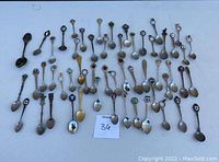 Overview of approximately 48 collectible souvenir spoons, laid out flat in rows for display. Spoons feature various metallic finishes with colorful enamel crests and unique handle designs.
