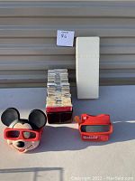 Photo showing two View-Master viewers: one shaped as Mickey Mouse and one standard red viewer, together with multiple slide packets and a white plastic slide storage case.