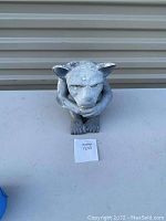 Front view of a small vintage gargoyle figurine sitting with chin on folded arms, showing weathered surface and detail of face and limbs.